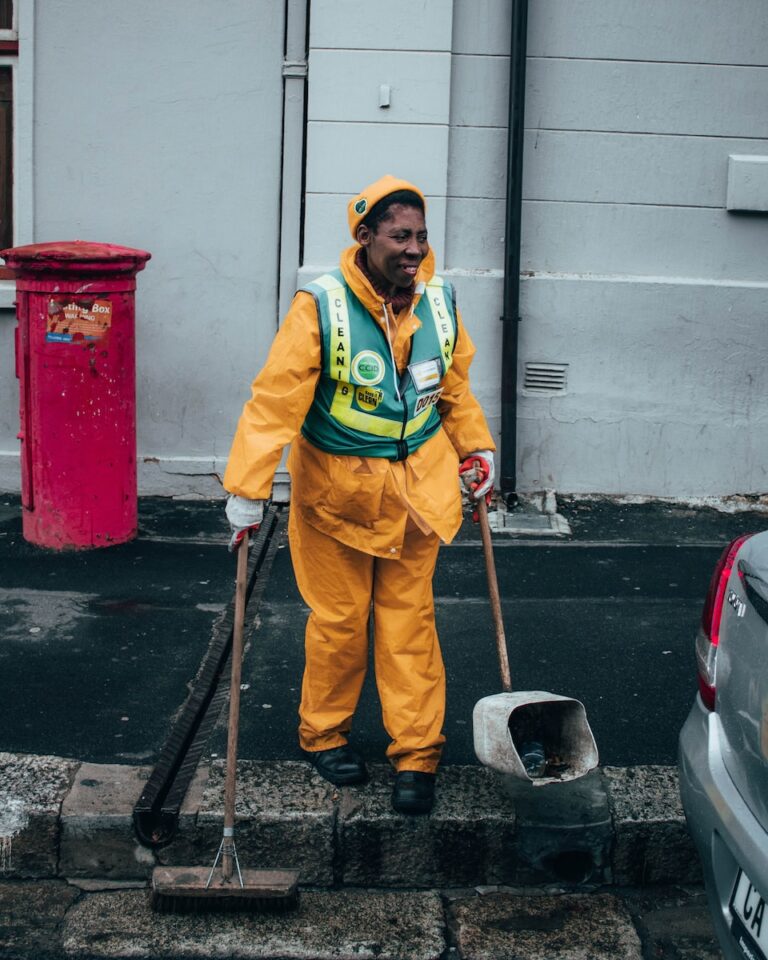 woman in green and yellow jacket holding black and brown stick standing beside red trash bin